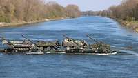 Vier Militärfahrzeuge stehen hintereinander auf einer Fähre und werden über den Fluss transportiert.