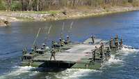 Eine leere Fähre mit mehreren Soldaten fährt einen Fluss entlang.