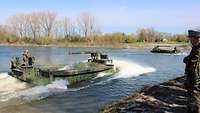 Zwei Amphibienfahrzeuge schwimmen auf der Donau. Ein Soldat steht am Ufer.