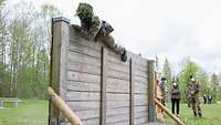 Ein Soldat überwindet eine zwei Meter hohe Holzwand. Zwei Soldaten und eine Zivilistin stehen am Rand und schauen zu.