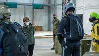 Eine Zivilistin steht vor zwei Soldaten in dunklen Freifallanzügen mit Helm. Rechts steht ein Springer mit gelbem Anzug.