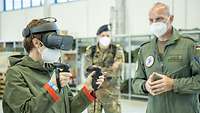 Eine Zivilistin trägt eine Virtual-Reality-Brille und hält zwei Controller in den Händen. Ein Soldat steht erklärend daneben.