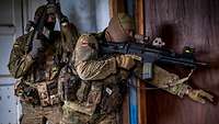 Zwei Soldaten stehen mit Waffen im Anschlag seitlich vor der Eingangstür eines Hauses und öffnen sie vorsichtig.