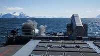 Blick auf das Vorschiff eines Kriegsschiffs in See: eine Kanone feuert nach links.