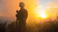 Ein Soldat steht in Uniform und mit Gewehr auf einem Übungsplatz im Abendrot.