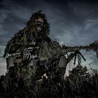 Ein Soldat in einem aufwendigen Tarnanzug kniet mit seinem Gewehr in der Dämmerung im Gelände. 