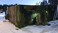 Two green containers on snow-covered ground. A man is standing in front of them. 