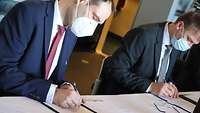 Two men seated at a table with distance, each signing a document