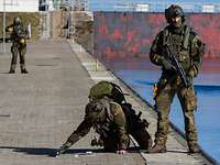 Zwei Soldaten in Flecktarnuniform auf ein Hafenpier; einer kniet und zeichnet etwas aufs Pflaster.