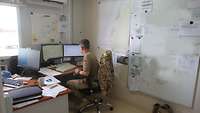 Ein Soldat sitzt in einem Büro am Computer. Die Wände hängen voll mit Skizzen, Notizen und Seekarten