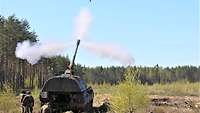 Eine Panzerhaubitze steht auf unebenem Gelände und feuert ein Geschoss ab, im Hintergrund Wald 