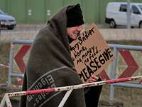 Ein Mann, gehüllt in eine große, braun-graue Wolldecke, hält ein Pappschild mit der Aufschrift: Please#en give#en.