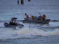 Ein kleines graues Boot mit Deckshäusern und Fenstern; davor ein dunkelgraues Speedboot mit großer, schaumiger Schraubenwelle.