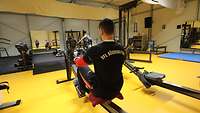 Ein Soldat sitzt in einem großen Fitnesszelt vor einem Spiegel auf einem Rudergerät und treibt Sport