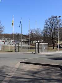 Gitterzaun und Gittertore, dahinter Fahnenmasten, davor ist das Wappen des Sanitätsregiments rechts vom Eingangstor angebracht