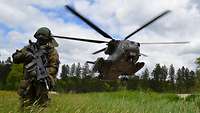 Auf einer Wiese kniet ein Soldat. Im Hintergrund landet ein Hubschrauber.