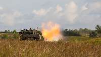 Ein Panzer steht im Gelände und schießt, ein Feuerball an der Kanonenmündung entsteht.