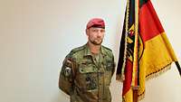 Ein Soldat steht vor einer weißen Wand und schaut in die Kamera, rechts von ihm die Truppenfahne.