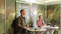 Ein Soldat in Flecktarn-Uniformund eine Frau in rosa Jacke stehen vor Mikrofonen. Zwischen ihnen eine Plexiglasscheibe.