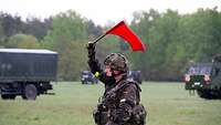 Ein Soldat schwenkt eine rote Flagge, im Hintergrund sind fahrende Fahrzeuge zu sehen.