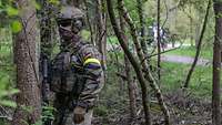 Ein vermummter Soldat mit Helm und Sonnenbrille steht in einem Wald.