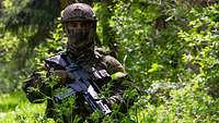 Ein vermummter Soldat mit Helm und G36 kniet in einem Gebüsch.