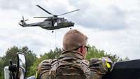 Ein Soldat lehnt im Vordergrund auf einem Auto. Im Hintergrund fliegt ein Hubschrauber.