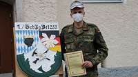 Ein Soldat steht vor einem Gebäude und hält eine Urkunde in Hand, neben ihm das Kompaniewappen.