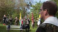 Eine Menschengruppe macht Rast im Grünen. Im Vordergrund ein Militärdekan in Uniform, im Hintergrund Bäume und die Flaggenträger