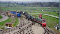 Ein roter Zug mit vielen Containern und Militärfahrzeugen fährt in einen Bahnhof ein