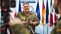 Ein Soldat steht vor Nationalflaggen und spricht in ein ihm hingehaltenes Mikrofon