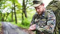 Ein Soldat steht auf einer Straße im Wald. Er trägt einen Rucksack und schaut auf seine Armbanduhr.