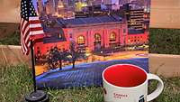Symbolfoto: ein amerikanischer Wimpel, ein Bild mit City-Skyline und Kaffeetasse mit Kansas-Aufdruck auf einer Wiese