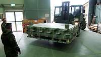 Die Transportpalette kann beladen werden Ein Gabelstapler transportiert eine Palette in eine Werkhalle. Davor stehen zwei Soldaten