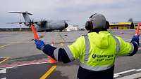 Ein Mitarbeiter der Flughafensicherheit steht mit vor dem Körper seitlich ausgestreckten Armen vor dem Flugzeug