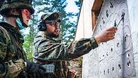 Zwei Soldaten stehen an einer Tafel mit Zielscheiben. Einer weist auf eine durchlöcherte Scheibe.
