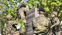Ein Soldat mit Waffe und Gepäck überwindet gerade eine Holzwand. Ein Soldat hilft.