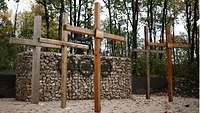 Fünf Holzkreuze stehen auf Sandboden vor einem Steinmonument, im Hintergrund ein Wald.