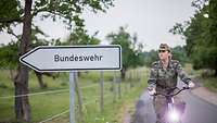 Eine Soldatin fährt mit einem Fahrrad an einem Wegweiser Bundeswehr vorbei.