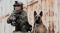 Ein Soldat in Kampfanzug hockt mit seinem Hund vor einer Fahrzeughalle.