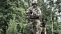Ein Soldat in Kampfanzug steht mit seinem Hund an einer Waldkante.