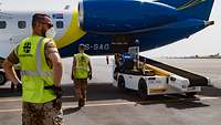Ein Soldat mit einer Warnweste steht vor einem zivilen Flugzeug, davor ein Wagen mit Gepäckband 