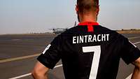 Ein Soldat steht bekleidet mit einem Fußballtrikot von Eintracht Frankfurt vor einem Flugfeld, im Hintergrund ein Flugzeug