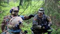 Ein belgischer Soldat zeigt ins Gelände. Ein deutscher Soldat kniet neben ihm und schaut in seine Richtung