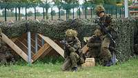Drei Soldaten stehen und hocken mit ihren Waffen vor einer mit Tarnnetz versehenen Holzkonstruktion.