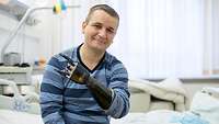 Ein Mann mit einer Armprothese sitzt auf einem Krankenhausbett und lächelt in die Kamera