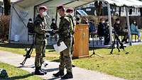 Ein Soldat reicht einem zweiten die Hand und gratuliert, im Hintergrund ein weißes Zelt mit Gästen auf Stühlen.