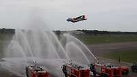 Drei Feuerwehrautos stehen am Rand der Landebahn und verschießen Wasser auf ein landendes Flugzeug.