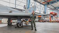 In a hangar, a man in an olive overall is standing in front of a gray aircraft