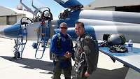 Two men standing in front of a jet aircraft, laughing and shaking hands